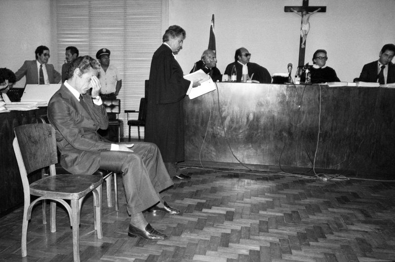 Foto do julgamento de Doca Street, assassino de Ângela Diniz. Imagem com Doca sentado à esquerda e outros homens em pé atrás. Fim da descrição