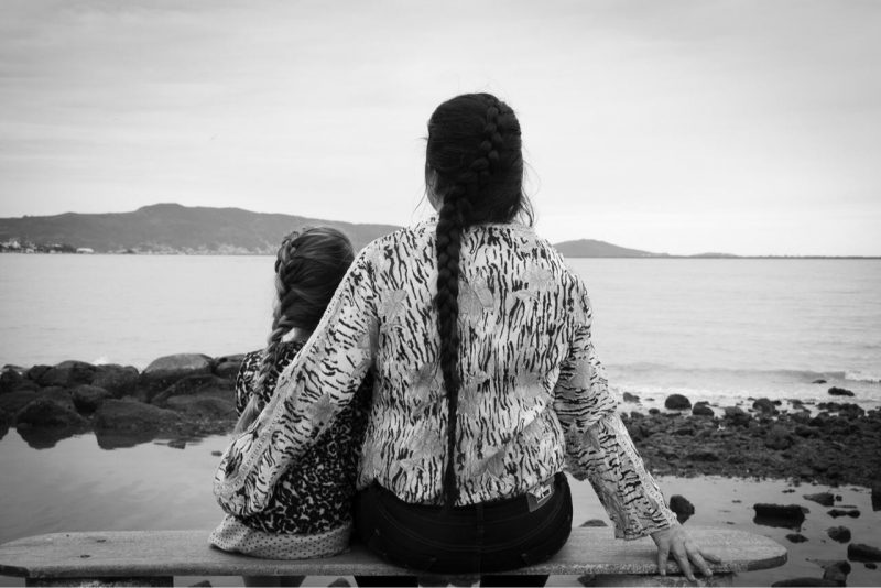 Foto de mãe e filha sentadas em um banco de frente pro mar. Fim da descrição